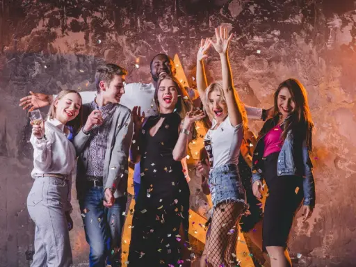 Grupa młodych ludzi świętuje, sypiąc konfetti. Ich stroje to świetna inspiracja, jak się ubrać na dyskotekę: sukienki, jeansy i błyszczące dodatki.