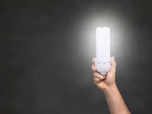 Dłoń trzymająca energooszczędną żarówkę o mocy 806 lumenów, rozświetlającą ciemne tło.