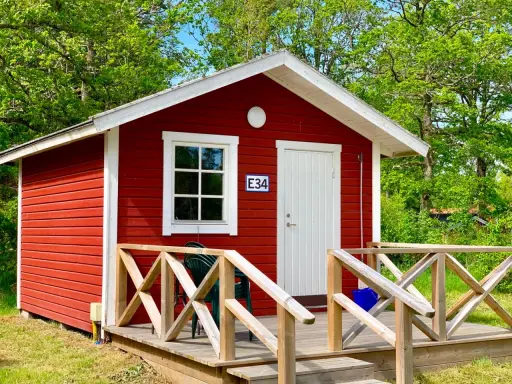 Rött litet hus med vit dörr och fönster, nummer E34. Trädäck med räcke. En grön stol står på däcket. Välkommen till Askeviks Camping!