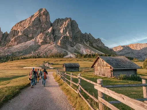 Piękna fotografia krajobrazowa Dolomitów. Grupa turystów wędruje ścieżką obok drewnianych chat, pod majestatycznymi szczytami górskimi.