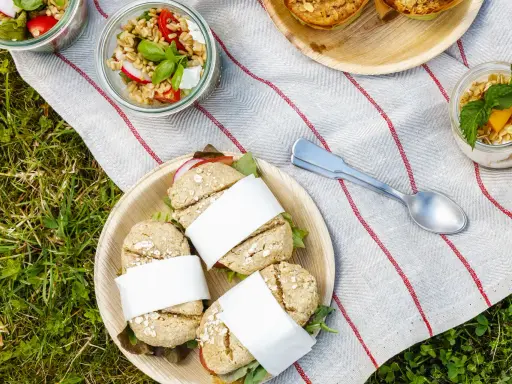 Idée pique-nique : petits pains garnis, salades en bocaux et muffins sur une nappe rayée, posée sur l'herbe.