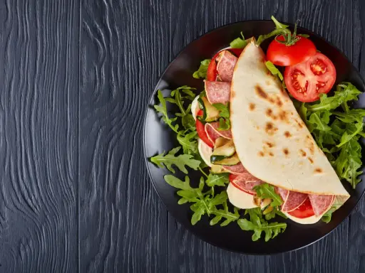 Piadina ripiena con salame, mozzarella, zucchine grigliate e pomodoro, servita su un piatto nero con rucola. Un assaggio della città di Rimini in un ristorante.
