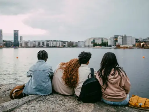 Three young people sit on rocks by the water, looking at a city skyline.