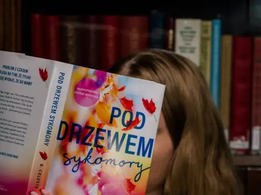 Dziewczyna czyta książkę "Pod drzewem sykomory", ukrywając twarz za okładką. W tle widać regały pełne książek.
