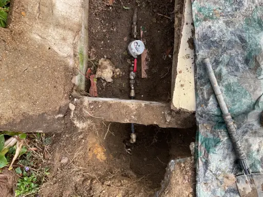 Réparation d'une fuite d'eau : un compteur et des tuyaux sont visibles dans une excavation, avec une bâche de protection à proximité.