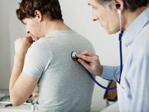 Arzt untersucht Patienten mit Stethoskop. Der Patient hustet, was auf Kurzatmigkeit hindeuten könnte.