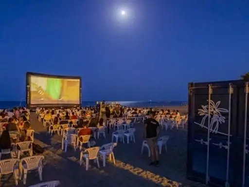 Noche de cine en el parque del cine Málaga. Público sentado en sillas en la playa, viendo una película en una pantalla gigante bajo la luna.