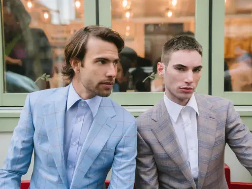 Two men in plaid suits with wide notch lapels sit side-by-side. The man on the left wears a light blue suit, the man on the right a tan suit.