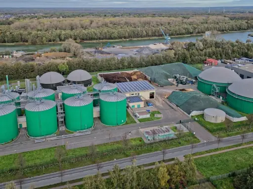 Grüne Biogas-Behälter und große Gasspeicher prägen diese Anlage, die die **Biogas Zukunft** symbolisiert. Im Hintergrund ein Fluss und bewaldete Ufer.