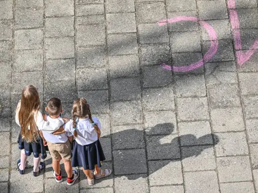 Trójka dzieci w szkolnych strojach patrzy na napis "szkoła społeczna co to" narysowany kredą.