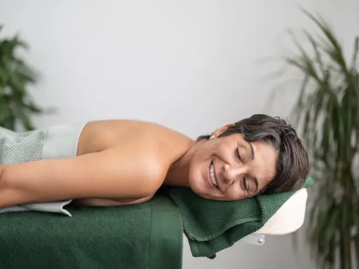 En kvinna ler och njuter av att vila på en massagebänk, insvept i en ljusgrön handduk. Gröna växter i bakgrunden bidrar till en lugn atmosfär.
