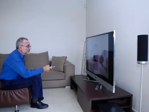 Starszy mężczyzna w okularach siedzi na kanapie i ogląda telewizję, trzymając pilota.