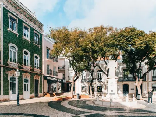 Grüne Kacheln zieren ein Gebäude in der Stadt Lagos. Ein Denkmal steht im Zentrum des Platzes, umgeben von Bäumen und Geschäften.