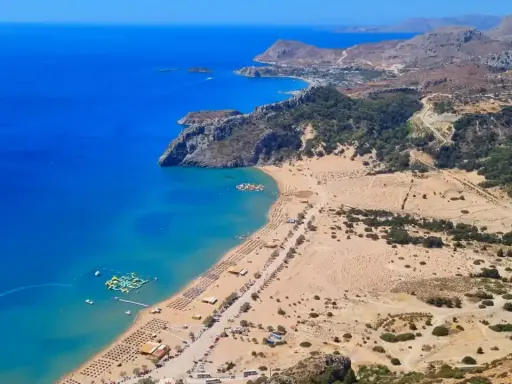 Rodos zachwyca turkusowym morzem i piaszczystą plażą. W oddali widać góry i zabudowania. Idealne miejsce na wakacje.