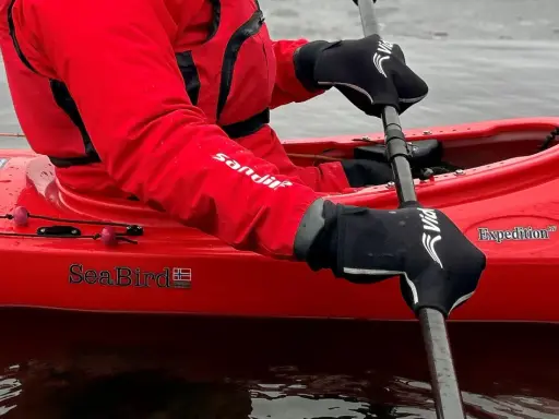 Person i röd jacka och svarta handskar paddlar kajak i kallt vatten.