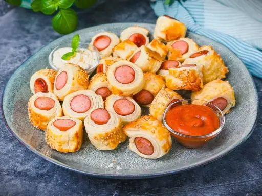 Leckeres Fingerfood für den Sommer: Würstchen im Blätterteig mit Sesam, serviert mit Dip und Ketchup.