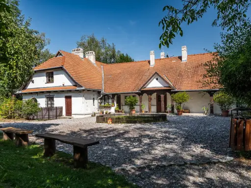Urokliwy dom z czerwonym dachem w Bronowicach Małych, otoczony zielenią. Widać drewniane ławki i kamienny dziedziniec.