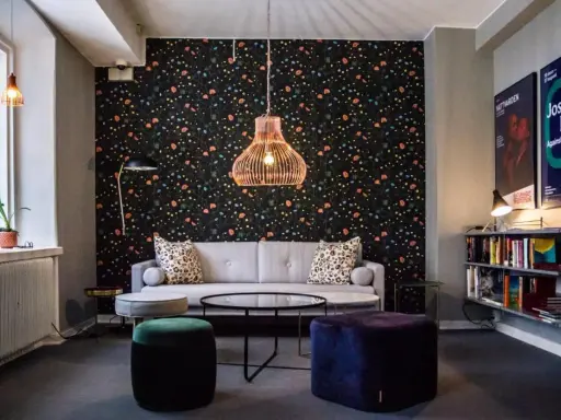 En mysig lounge med blommig tapet, en grå soffa, färgglada sittpuffar och en hylla full med böcker. En affisch för "Josef Frank - Against Design" hänger på väggen.