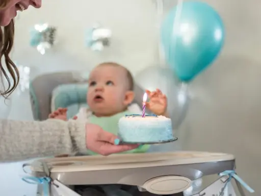 Mama podaje tort z jedną świeczką na roczek maluszkowi. Balony i dekoracje tworzą radosną atmosferę.