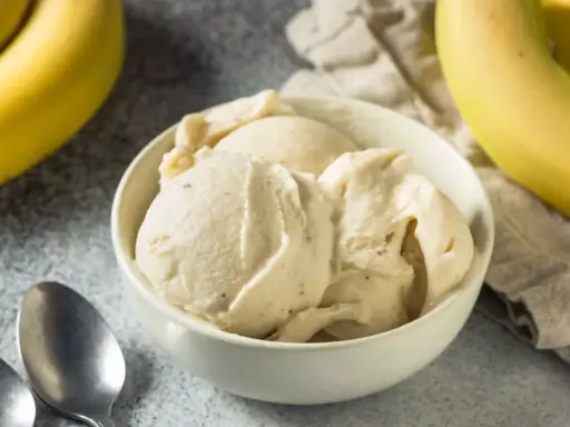 Un cremoso helado de platano servido en un cuenco blanco, con plátanos amarillos frescos al fondo.