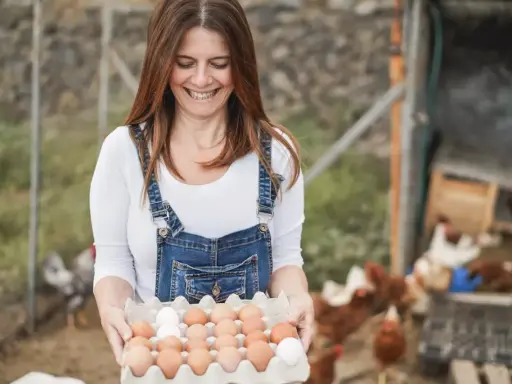 Uśmiechnięta kobieta w ogrodniczkach trzyma tacę z jajkami. To świetne sprzedaż bezpośrednia przykłady świeżych produktów prosto od kury.