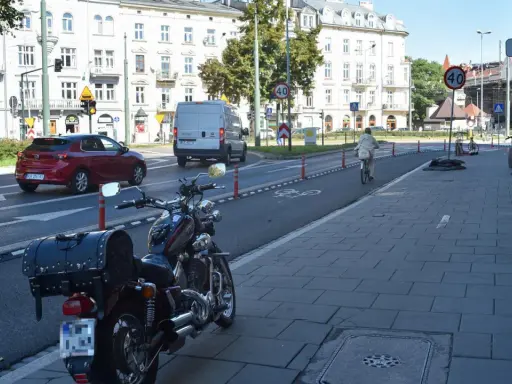 Motocykl zaparkowany na chodniku, obok drogi z wyznaczonym pasem rowerowym. W tle budynki i ruch uliczny, w tym czerwony samochód i biały van.