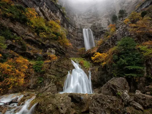 Cascada en un cañón rocoso, con árboles otoñales y niebla. El agua cae como un zafiro lagunazo, creando un espectáculo natural.