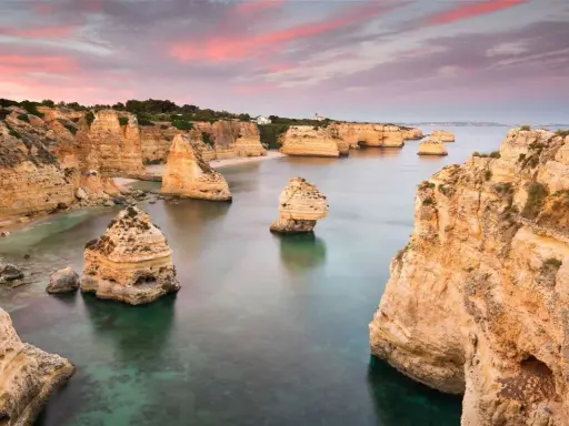 Felsformationen ragen aus dem ruhigen Wasser der **Lagoa, Portugal**, empor. Der Himmel ist in Rosa- und Orangetönen gefärbt, was auf einen wunderschönen Sonnenuntergang hindeutet.