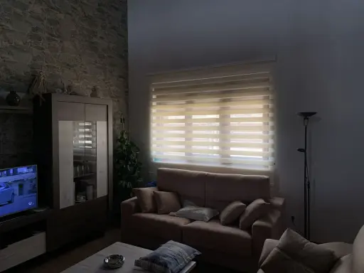 Salón acogedor con estores enrollables noche y día que filtran la luz, sofá, mueble y televisión.