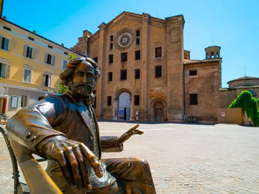 Statua bronzea in una piazza di una città dell'Emilia Romagna, con un'antica chiesa in mattoni sullo sfondo.