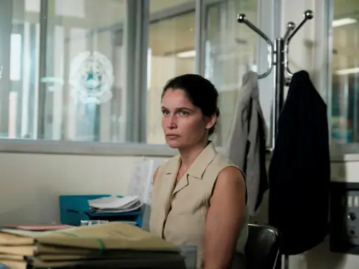 Una donna con un'espressione pensierosa siede in un ufficio disordinato, circondata da fascicoli. Sembra l'inizio di una storia nera.