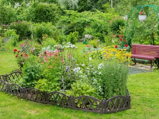 Un exuberante macizo de flores de verano, con rosas rojas, margaritas blancas y flores amarillas vibrantes, bordeado por una valla decorativa.