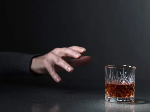 Mano tesa verso un bicchiere di whisky, un'immagine che evoca la domanda: quanto beve un alcolista al giorno?