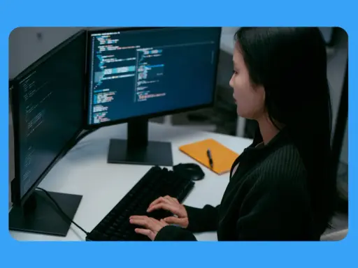 Jeune femme travaillant sur un ordinateur, avec deux écrans affichant du code. Sa position devant l'écran du bureau est concentrée.