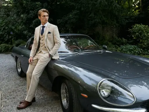 A man in a light-colored suit, perfect for mens summer cocktail attire, leans against a vintage car.