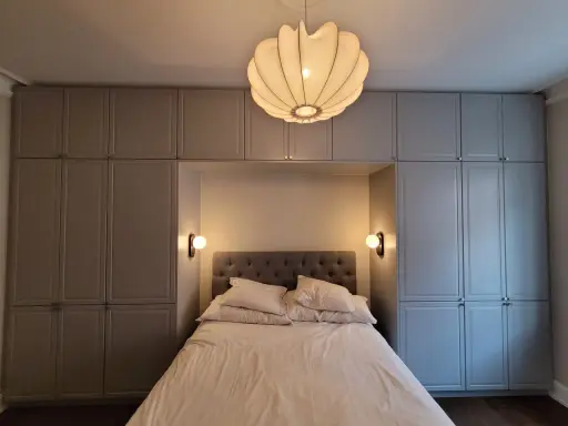 Sovrum med inbyggda garderober i en sofistikerad stil. En stor, mjuk säng med många kuddar står centrerad, belyst av en unik taklampa och två vägglampetter.