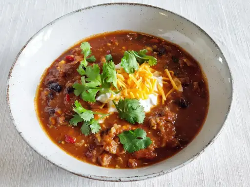 Gęste, aromatyczne chili con carne z fasolą, mięsem i przyprawami, udekorowane śmietaną, startym serem i świeżą kolendrą.