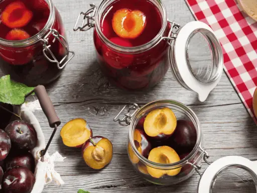 Frische Pflaumen werden für das Einkochen vorbereitet. Ganze und halbierte Früchte liegen neben Gläsern mit eingelegten Pflaumen.