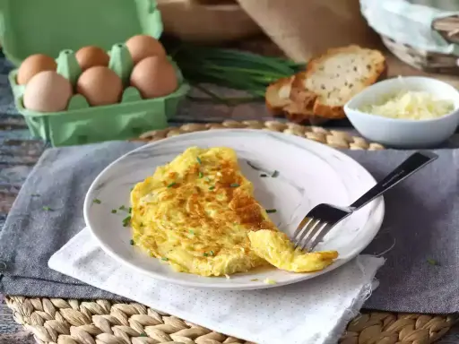 En gyllene omelett med ost, toppad med gräslök. Bredvid syns ägg, rostat bröd och mer ost. En perfekt start på dagen!