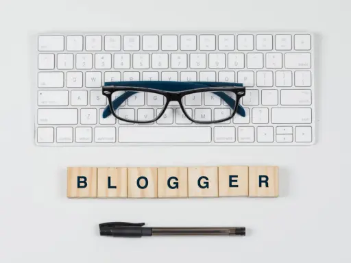 Okulary, klawiatura i drewniane klocki z napisem "BLOGGER" - idealne narzędzia do pisania bloga po angielsku.
