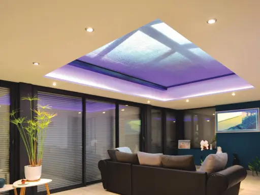 Salon moderne avec extension de maison et son puits de lumière illuminé de violet. Canapé confortable, plantes vertes et grandes baies vitrées.
