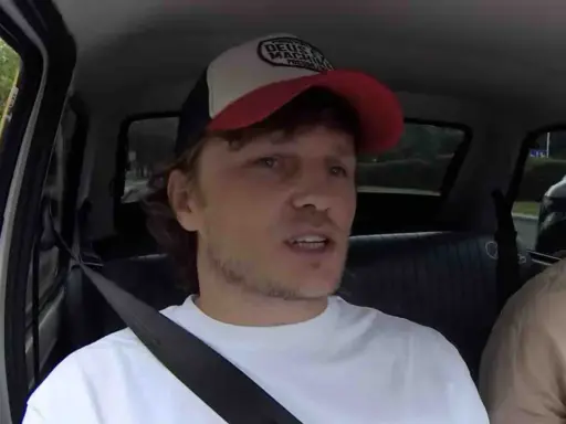 Mężczyzna w czapce z daszkiem i białej koszulce, siedzący w samochodzie. W tle widać drzewa i inne pojazdy. To może być początek jego zaorski wiek.