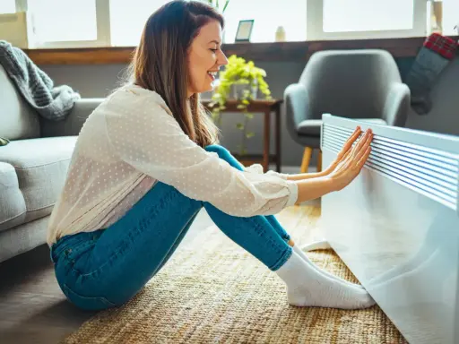 Mujer sonríe mientras calienta sus manos en un radiador, disfrutando de la temperatura ideal en casa en invierno.