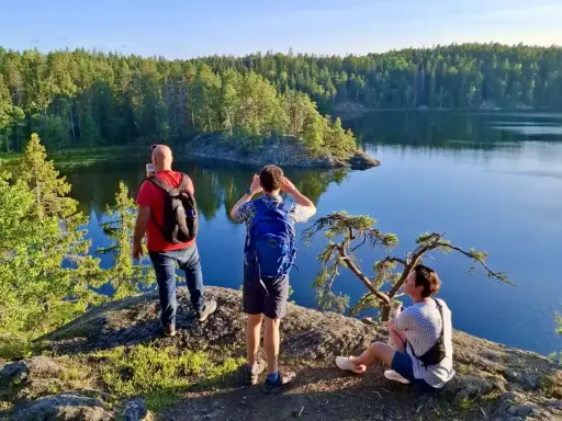 Vandring i Tyresta nationalpark. Tre personer njuter av utsikten över en stilla sjö och en tät skog. En man tar foto, en annan spanar ut, och en tredje vilar på en klippa.