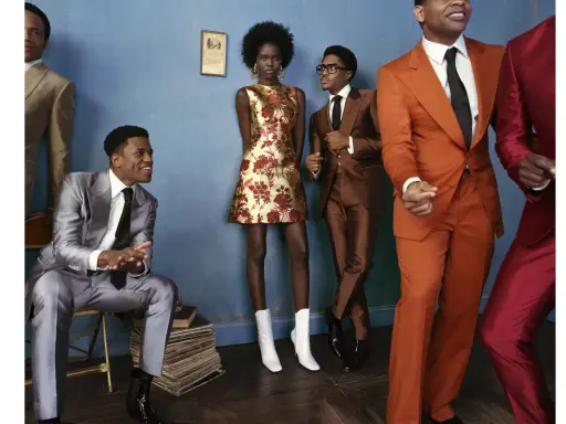 A group of stylish individuals in smart cocktail attire, featuring bold suits and a patterned dress, pose against a blue wall.
