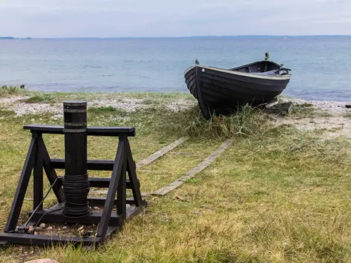 A dark wooden boat rests on a grassy shore next to a winch and wooden tracks leading to the sea.