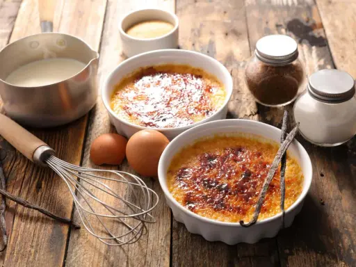 Dwa kremowe desery creme brulee z karmelizowanym cukrem, obok składniki: śmietanka, jajka, laski wanilii, cukier.
