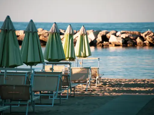 Ombrelloni verdi e lettini vuoti sulla spiaggia di una città riviera romagnola, con il mare calmo e una diga di sassi sullo sfondo.