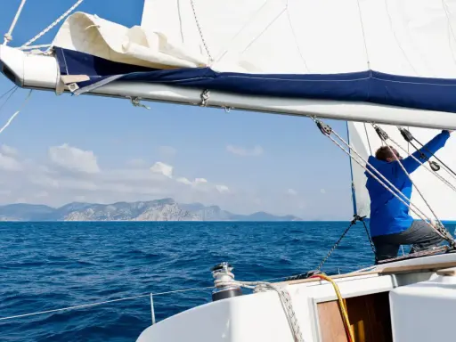 Mann im blauen Anzug stellt das **Rigg segeln** ein, während das Boot über das blaue Meer gleitet. Berge im Hintergrund.