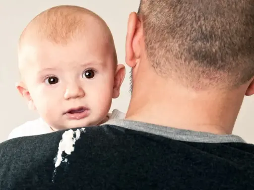 En söt bebis med stora ögon tittar fram över pappans axel. En liten vit fläck syns på pappans tröja, kanske från en rapning.